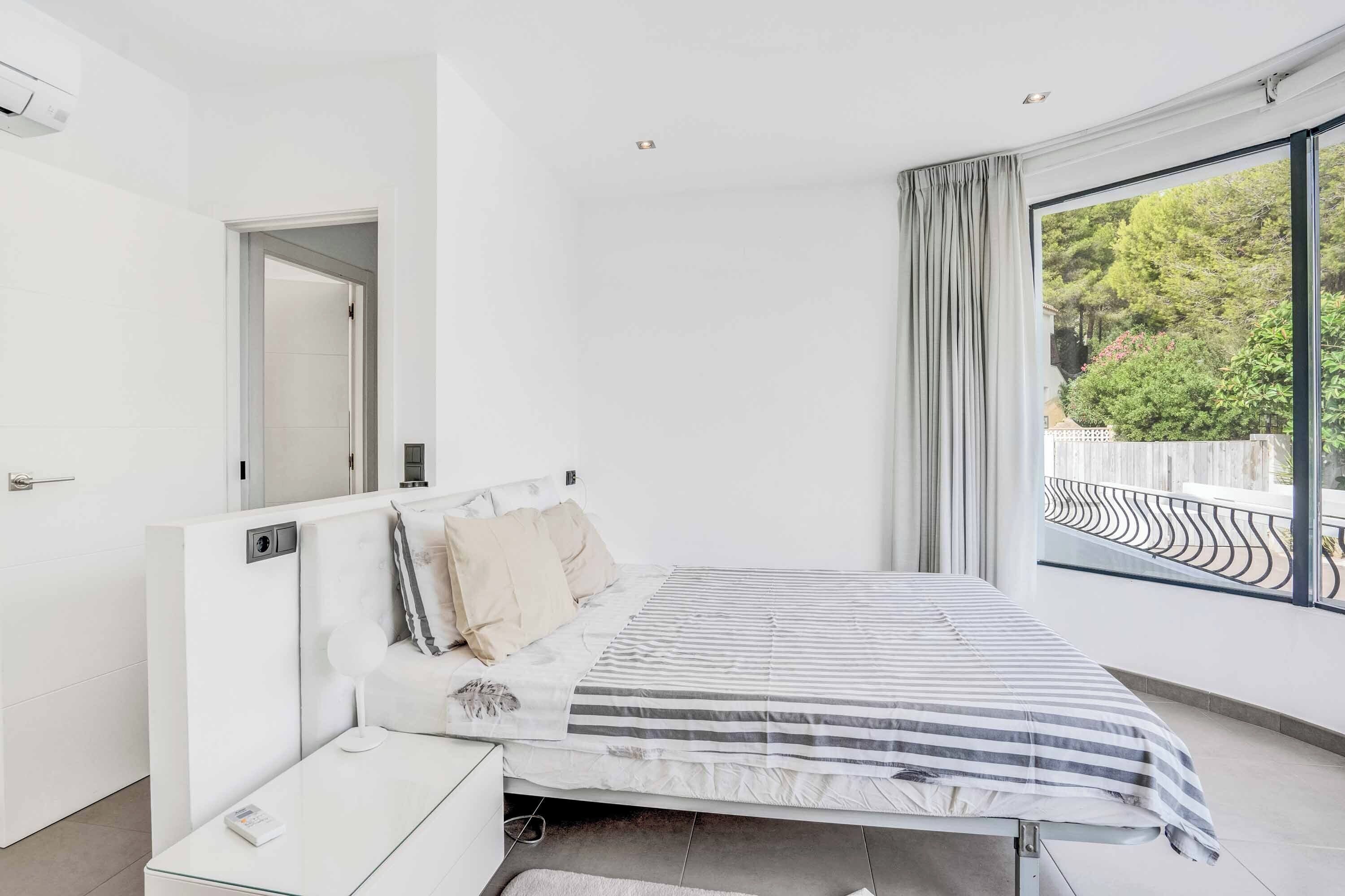 A modern bedroom with a large window, a bed with striped bedding, a bedside table, and a view of greenery outside.