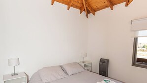 A bedroom with a wooden ceiling, a bed with a grey bedspread, two white bedside tables with lamps, and a window with a view.