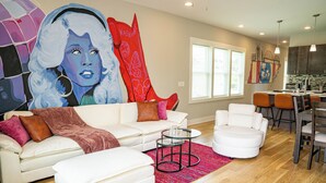A modern living room with a white sofa, a glass coffee table, and a colorful rug. There is a kitchen area with a dining table and chairs visible in the background.