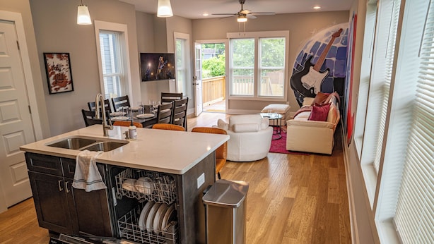 A modern kitchen with a dishwasher, a dining area with chairs, and a living room with a couch and a guitar.