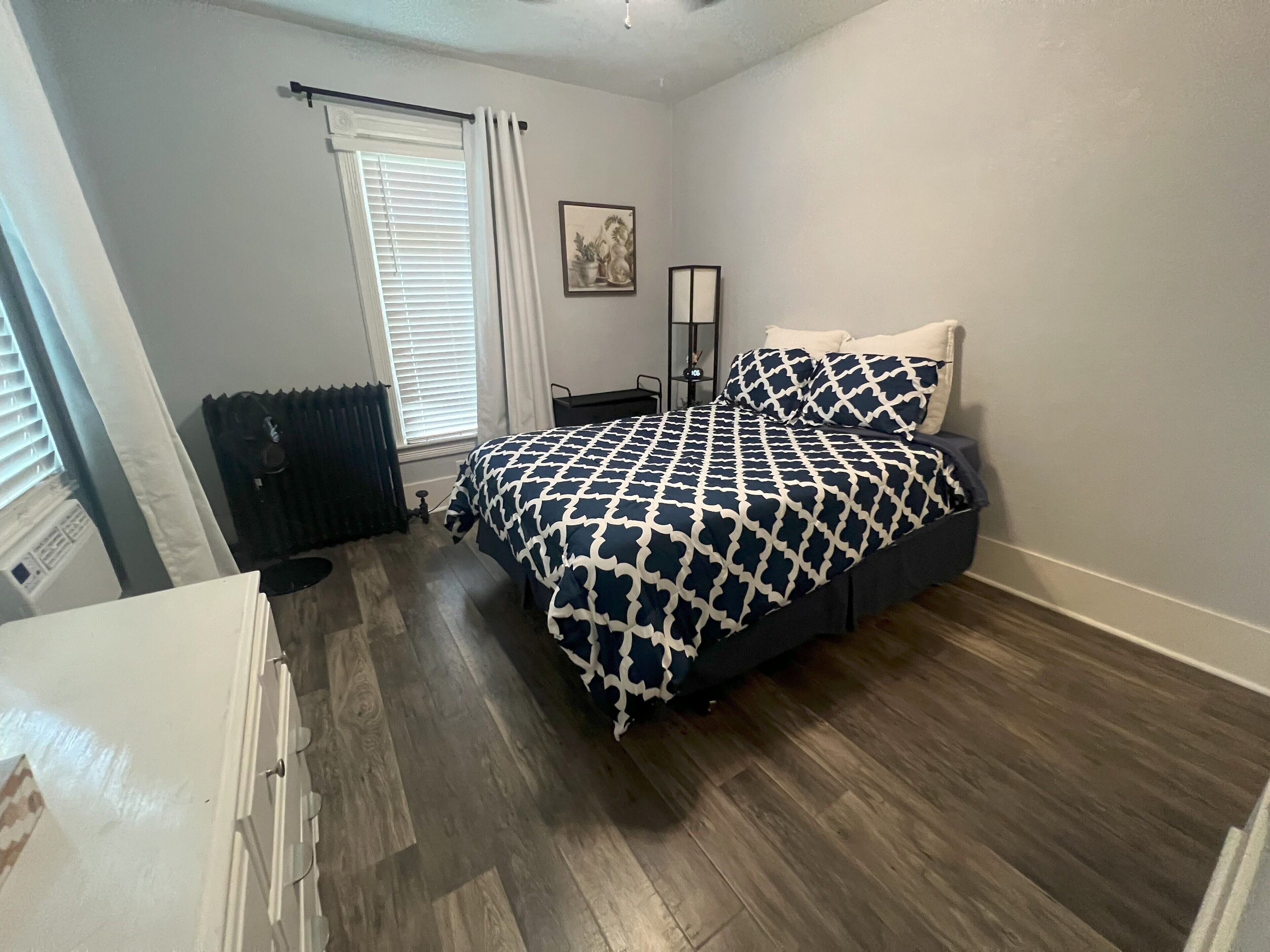 A bedroom with a bed, a radiator, a window with blinds, and a floor lamp.