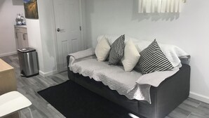 Ett litet, modernt vardagsrum med en grå soffa, en svart papperskorg, en vit dörr och en vit luftkonditioneringsenhet på väggen.