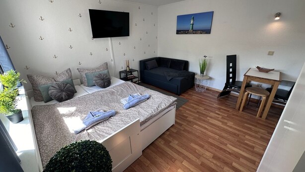Ein modernes, kompakter Wohnraum mit Bett, Sofa, kleinem Tisch und Wandfernseher.