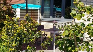 Een terras buiten met een tafel en stoelen, een roze parasol, en een raam met uitzicht op de straat.