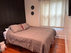 Quarto com cama, janela com cortinas, um respiradouro na parede e uma lixeira.