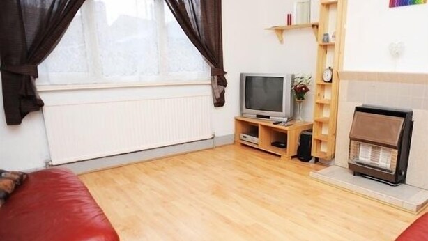 A living room with a wooden floor, a red sofa, a television, a small wooden shelf, and a radiator. - House - Sleeps 5, Private Garden & Free Parking (London)
