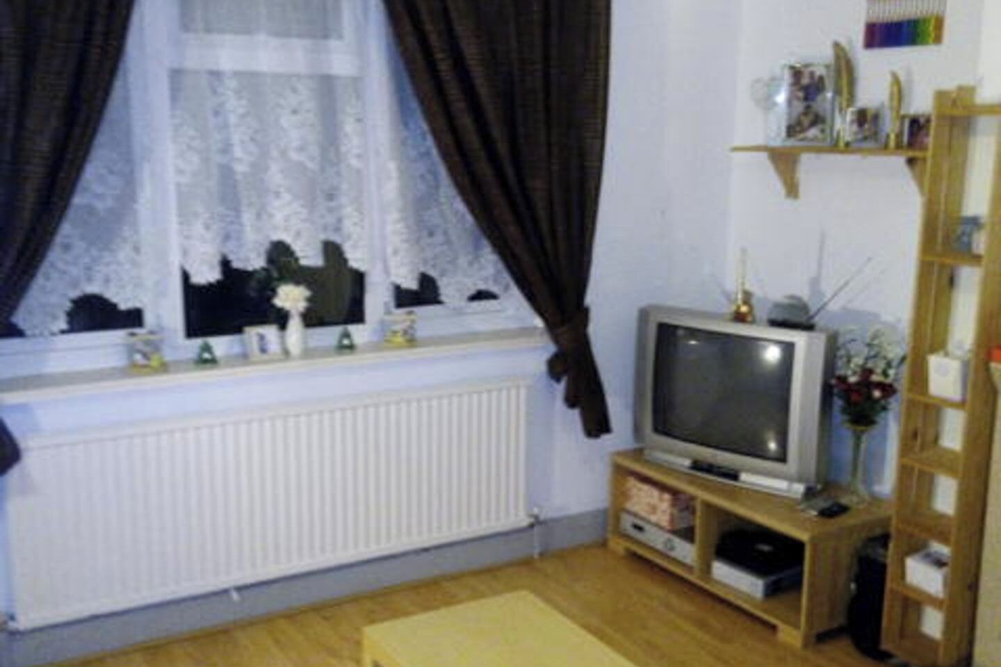 Une pièce avec un radiateur, un téléviseur posé sur un meuble en bois, une fenêtre avec des rideaux et une étagère contenant divers objets.