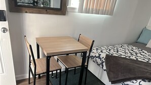Une pièce de taille modeste, avec une table et des chaises en bois, un lit avec une literie à motifs, une petite kitchenette et une fenêtre équipée de stores.