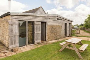 A rustic wooden building with a large sliding glass door, a stone wall, and a picnic table in the yard. - Cotswold Converted Milk Parlour (Stroud)