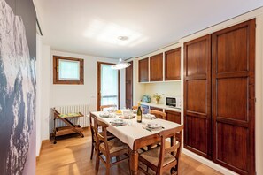 Un comedor con una mesa de madera preparada para cuatro personas, rodeada de sillas de madera. Hay un radiador en la pared y una ventana sobre la mesa.