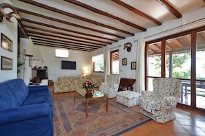 A living room with a blue sofa, patterned armchairs, a coffee table with a flower vase, and a large window with a view of greenery.