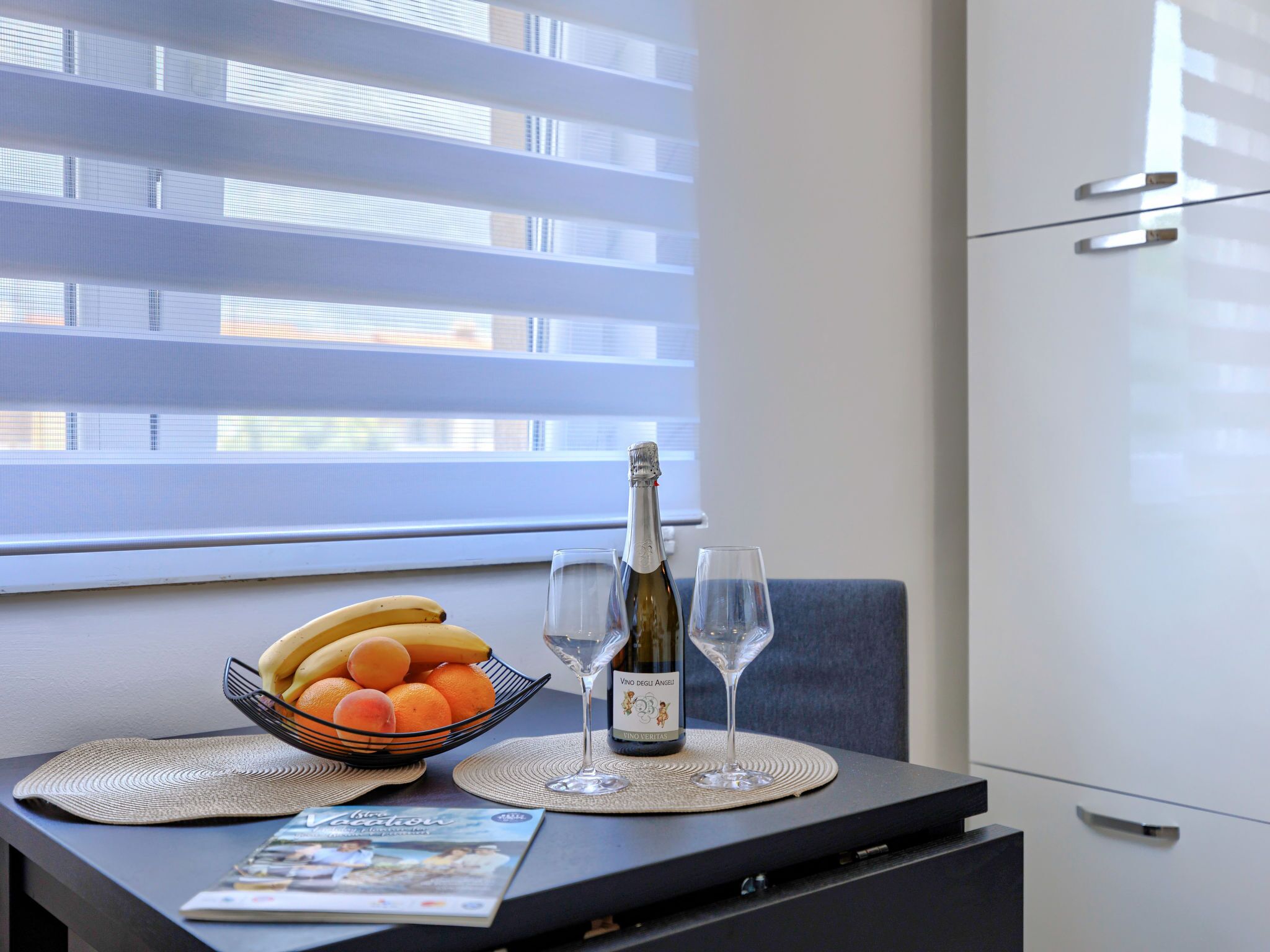 A dining table with a bottle of wine, two glasses, a bowl of fruit, and a magazine.