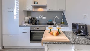 Une cuisine moderne avec des meubles blancs, un four encastré, une machine à café, un grille-pain et une planche à découper en bois sur laquelle sont disposés des fruits.