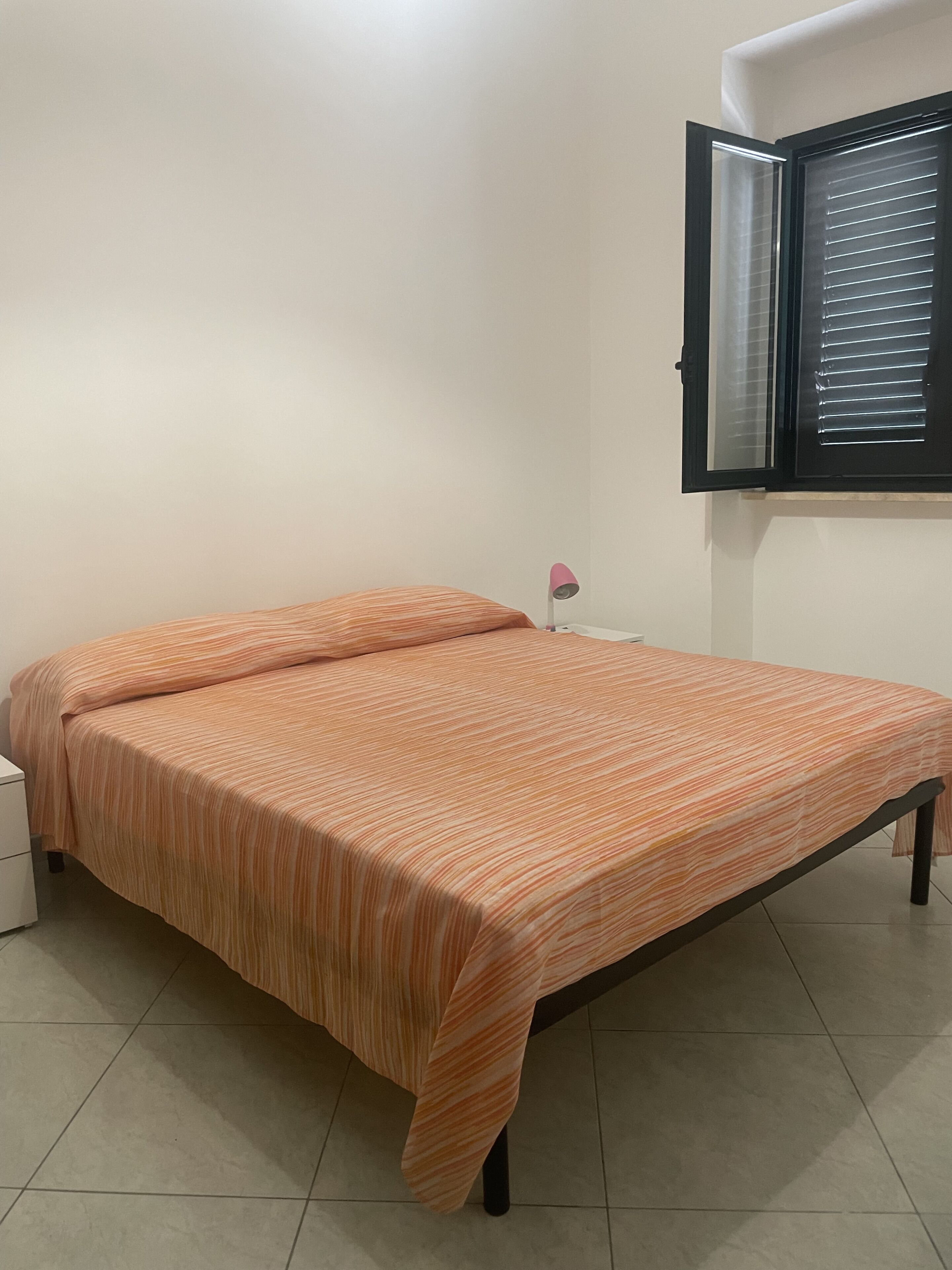 Un lit simple avec une couvre-lit à rayures, une table de chevet avec une lampe et une fenêtre avec des stores.