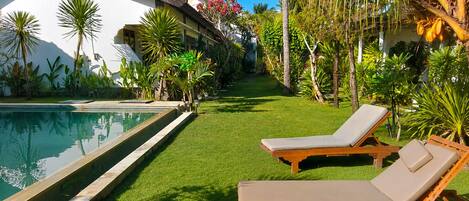 Un área junto a la piscina con un edificio blanco, palmeras y dos sillas de madera para relajarse.
