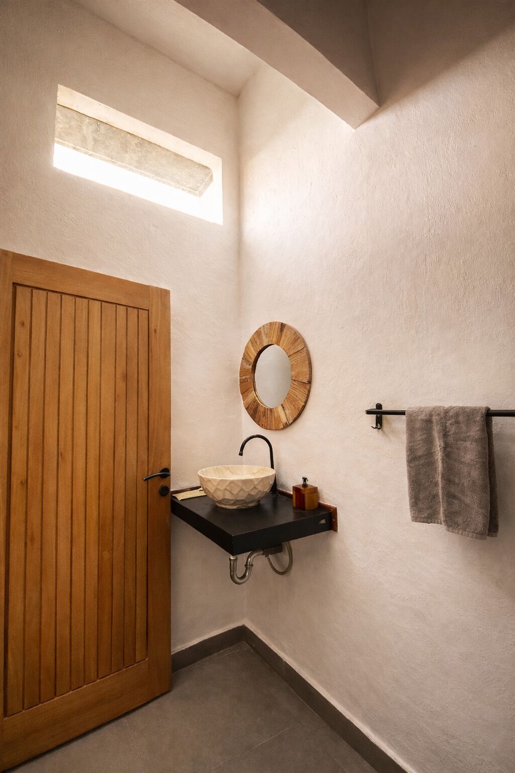 A bathroom with a wooden door, a round mirror, a sink with a black countertop, and a towel hanging on a rack.