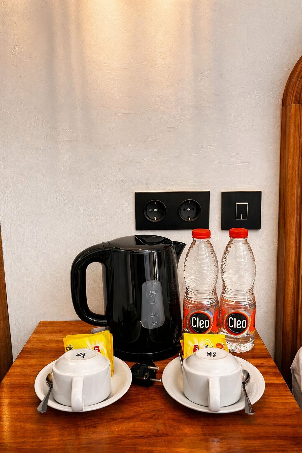 Un tavolo con un bollitore elettrico nero, due bottiglie di acqua Cleo e due tazze bianche con piattini.