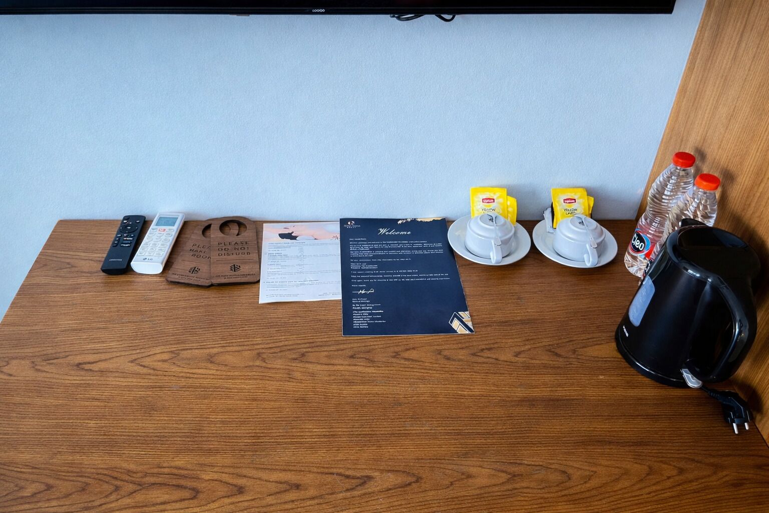 A wooden desk with a remote control, a wooden tray with a note, a welcome card, two yellow cups, a water bottle, and a black coffee maker.