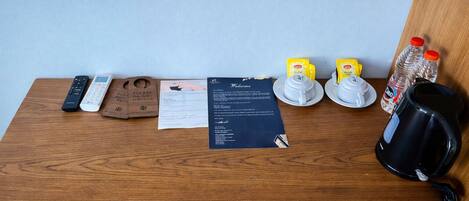 A wooden desk with a remote control, a wooden tray with a note, a welcome card, two yellow cups, a water bottle, and a black coffee maker.