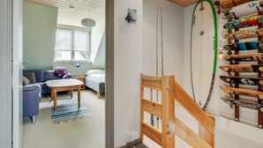 A room with a bed, a desk, and a window. There is a wooden staircase leading to a loft area with a surfboard rack.