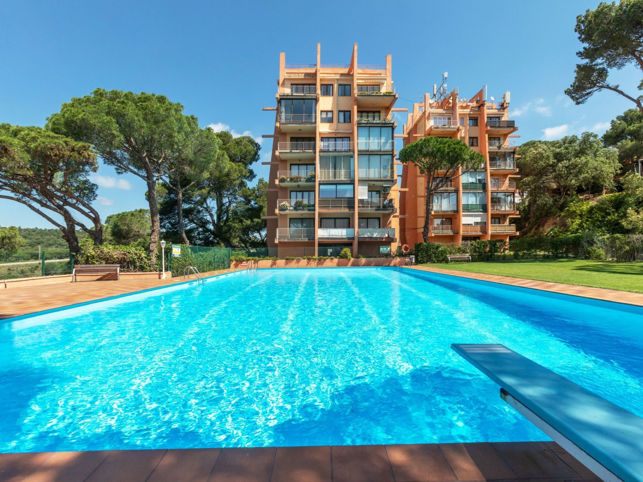 A modern hotel with a large swimming pool, surrounded by lush greenery and a clear blue sky.