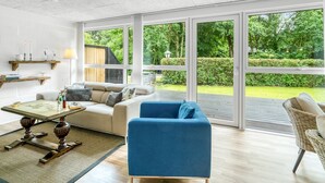 A spacious living room with a large sliding glass door leading to a garden, a sofa, a blue armchair, and a coffee table.
