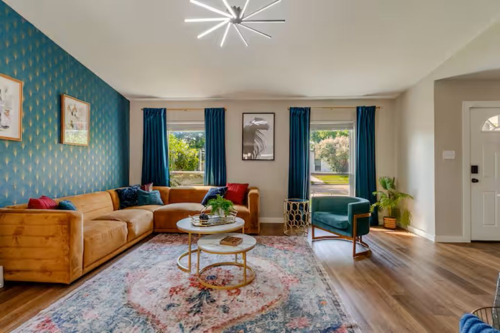 A living room with a sofa, a coffee table, and a chair. There are curtains on the windows, and a plant is visible.
