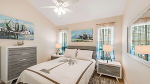A hotel room with a bed, a dresser, two nightstands with lamps, a ceiling fan, and two paintings on the walls. - Uncapafew more Beachside - Unit 1602 Island Dunes Condos- One block from Beach (Port Aransas)