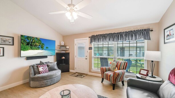 A living room with a grey sofa, a striped armchair, a television, a ceiling fan, and a window with blinds. - Uncapafew more Beachside - Unit 1602 Island Dunes Condos- One block from Beach (Port Aransas)