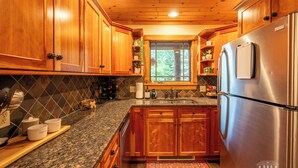 A kitchen with wooden cabinets, granite countertops, a stainless steel refrigerator, and a window with a view of greenery. - Mountain Retreat - walk to Main Street (Lake Placid)