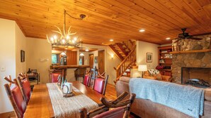 A spacious dining area with a wooden table and chairs, a stone fireplace, and a cozy living area with a couch and a staircase. - Mountain Retreat - walk to Main Street (Lake Placid)