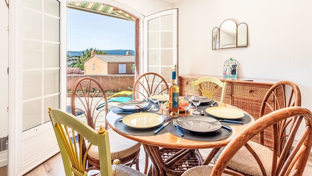 A dining area with a table set for four, a wicker chair, and a view of a house and landscape through open doors. - Les Mas de Cogolin by Interhome (Cogolin)