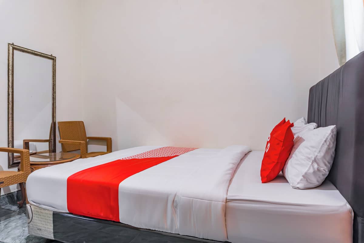 Un lit bien fait, avec une literie blanche rehaussée d’une touche de rouge, une chaise en bois et un miroir accroché au mur.