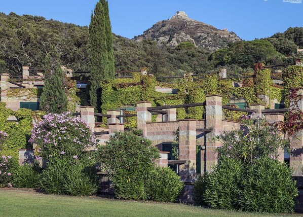 A building with stone walls, green shutters, and ivy-covered structures, set against a mountainous backdrop.