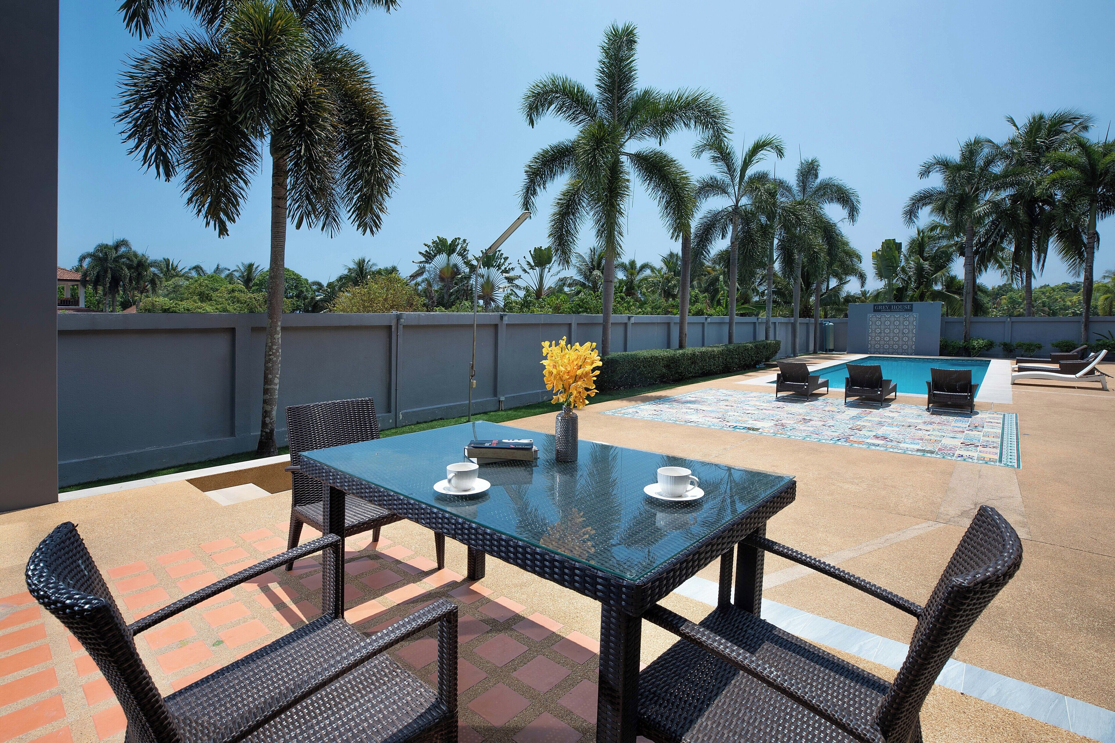 Eine Terrasse mit Glastisch und Korbstühlen, ein Pool und im Hintergrund Palmen.
