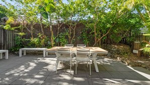 Une terrasse extérieure avec une table en bois et des chaises blanches, entourée d’une végétation luxuriante et d’un mur de pierre.