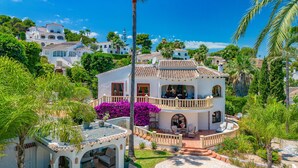Une grande maison à deux étages, dotée d’un balcon, entourée d’une végétation luxuriante et agrémentée d’une piscine.