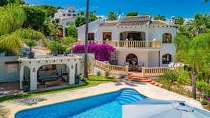 Une maison à deux étages avec une piscine, entourée d’une végétation luxuriante et de palmiers.