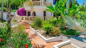 Une maison à deux étages avec un balcon, entourée de plantes tropicales et d’un jardin.