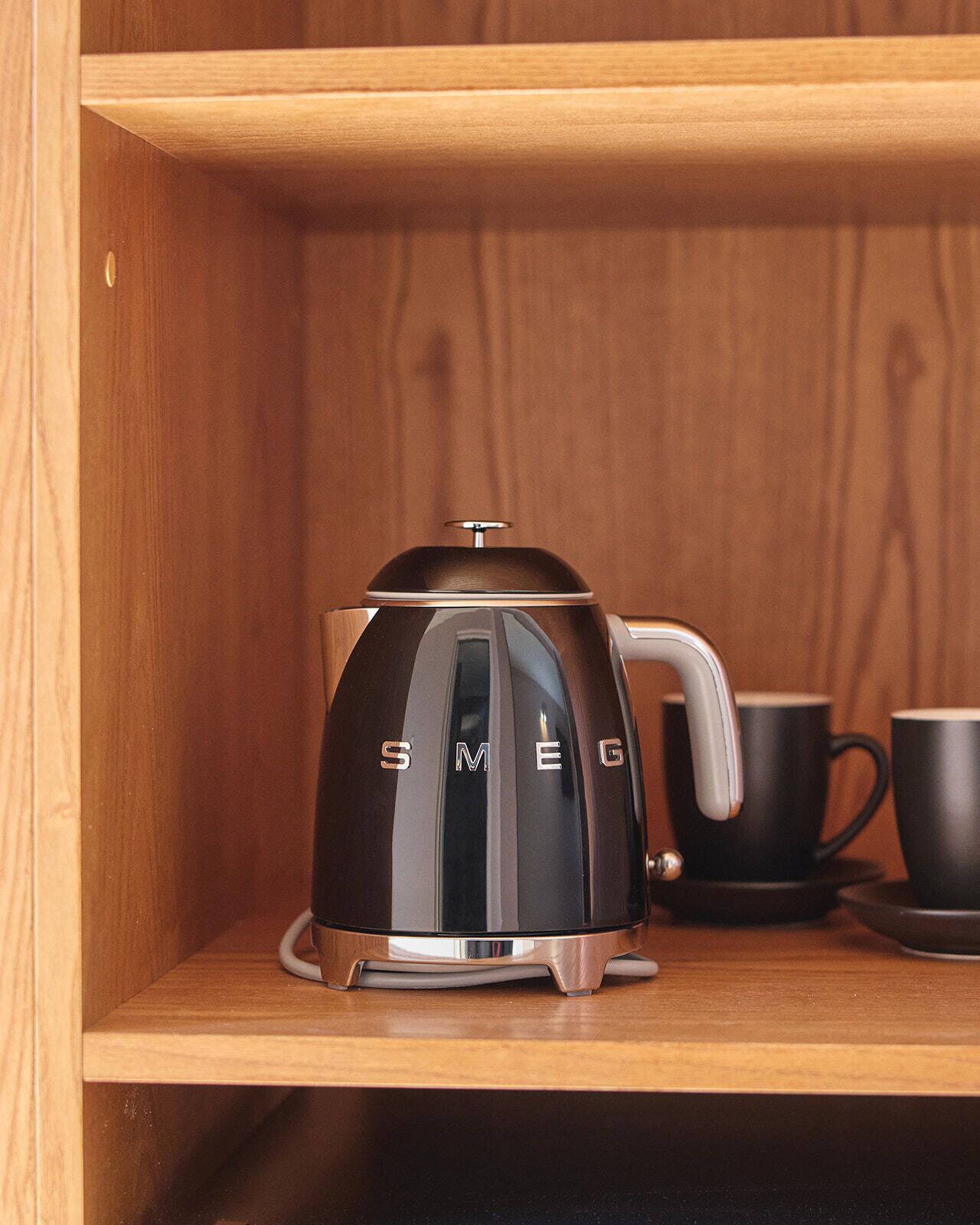 A black Smeg coffee pot on a wooden shelf with two black cups beside it.