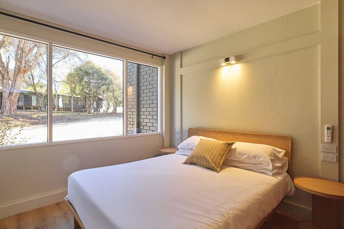Una cama bien hecha con cabecera de madera, mesita de noche y lámpara fijada a la pared.