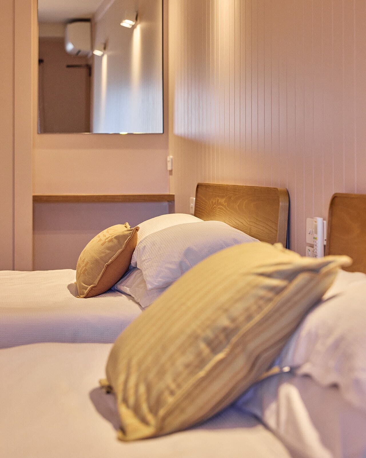 A hotel room with a bed, pillows, a wooden headboard, a wall-mounted mirror, and a bedside table.