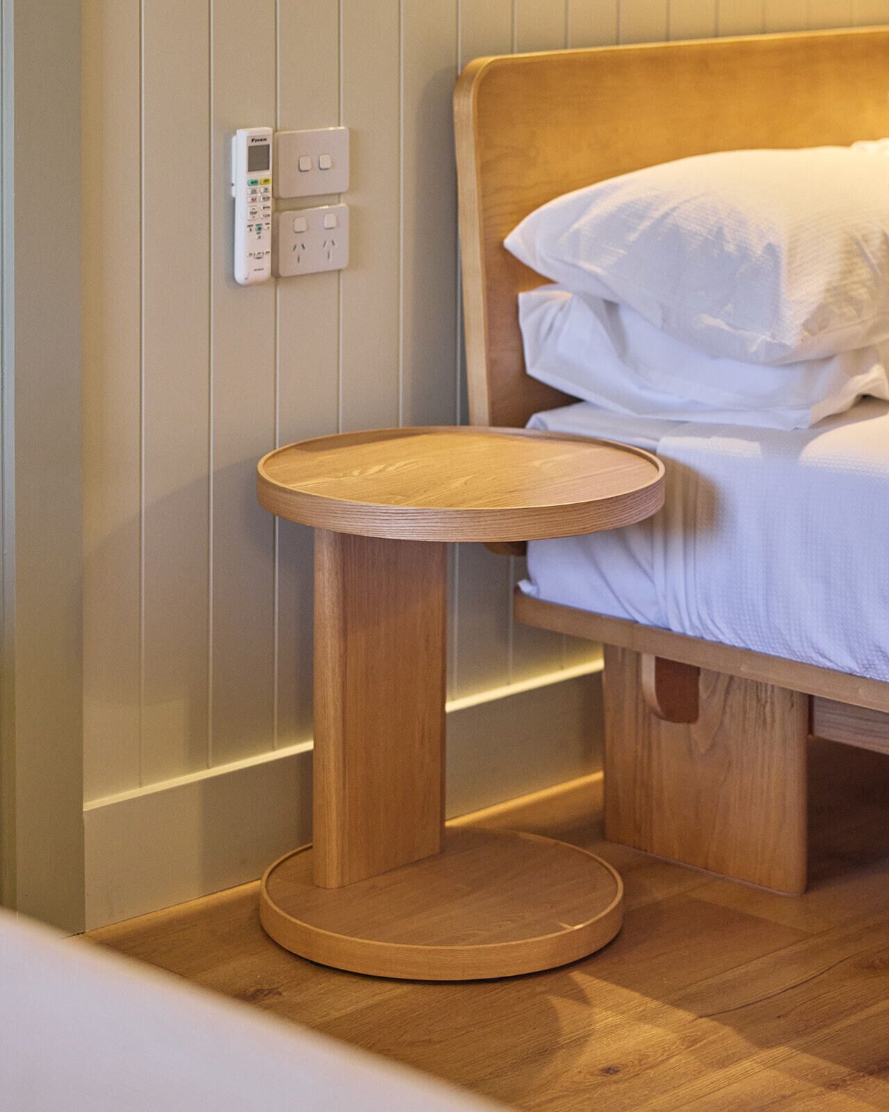 Een houten nachtkastje met een rond blad, geplaatst naast een bed met witte lakens.