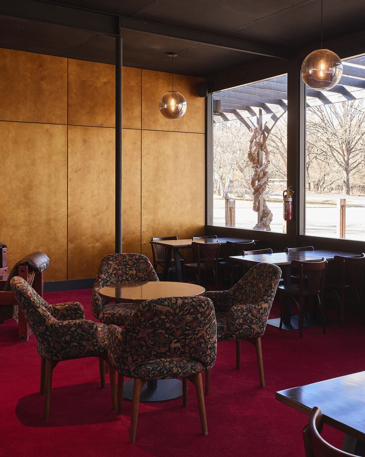 A restaurant interior with round tables and patterned chairs, a red carpet, and a view of trees through large windows.