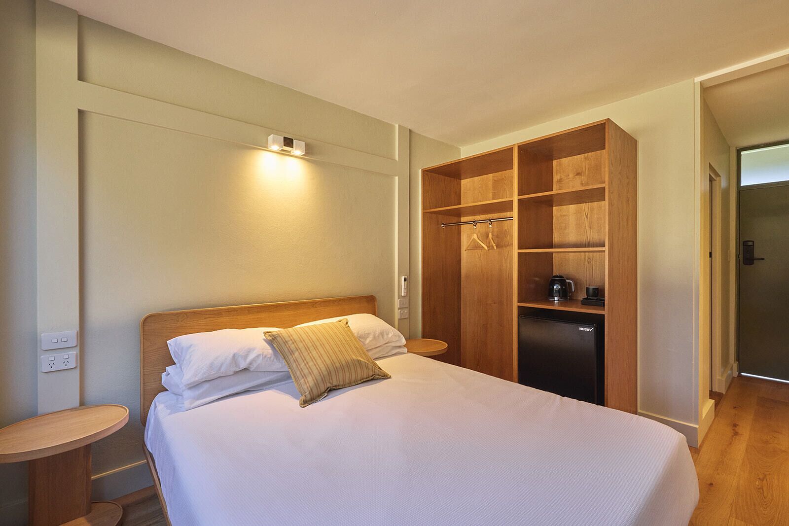 A neatly made bed with white linens and a striped pillow, a wooden wardrobe with a coffee maker inside, and a small bedside table.