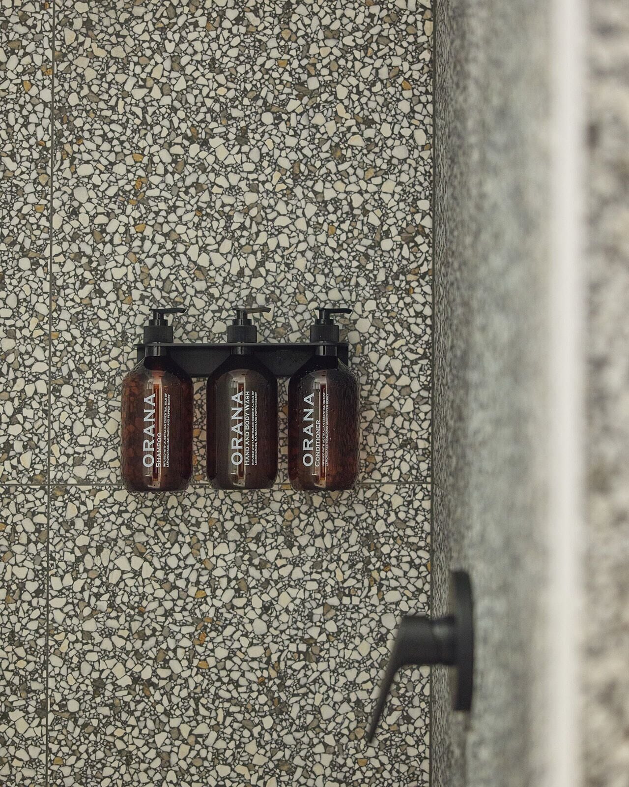 A bathroom with a terrazzo floor and three bottles of Orana hand and body wash on a black soap dispenser holder.