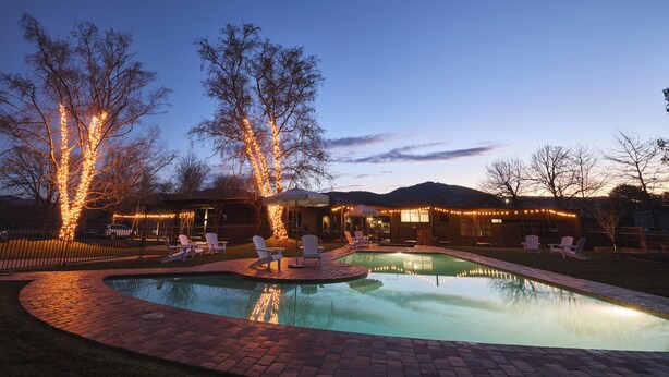A swimming pool with a brick deck, surrounded by lounge chairs and umbrellas, with string lights on trees and a house in the background. - Alpine Inn (Khancoban)