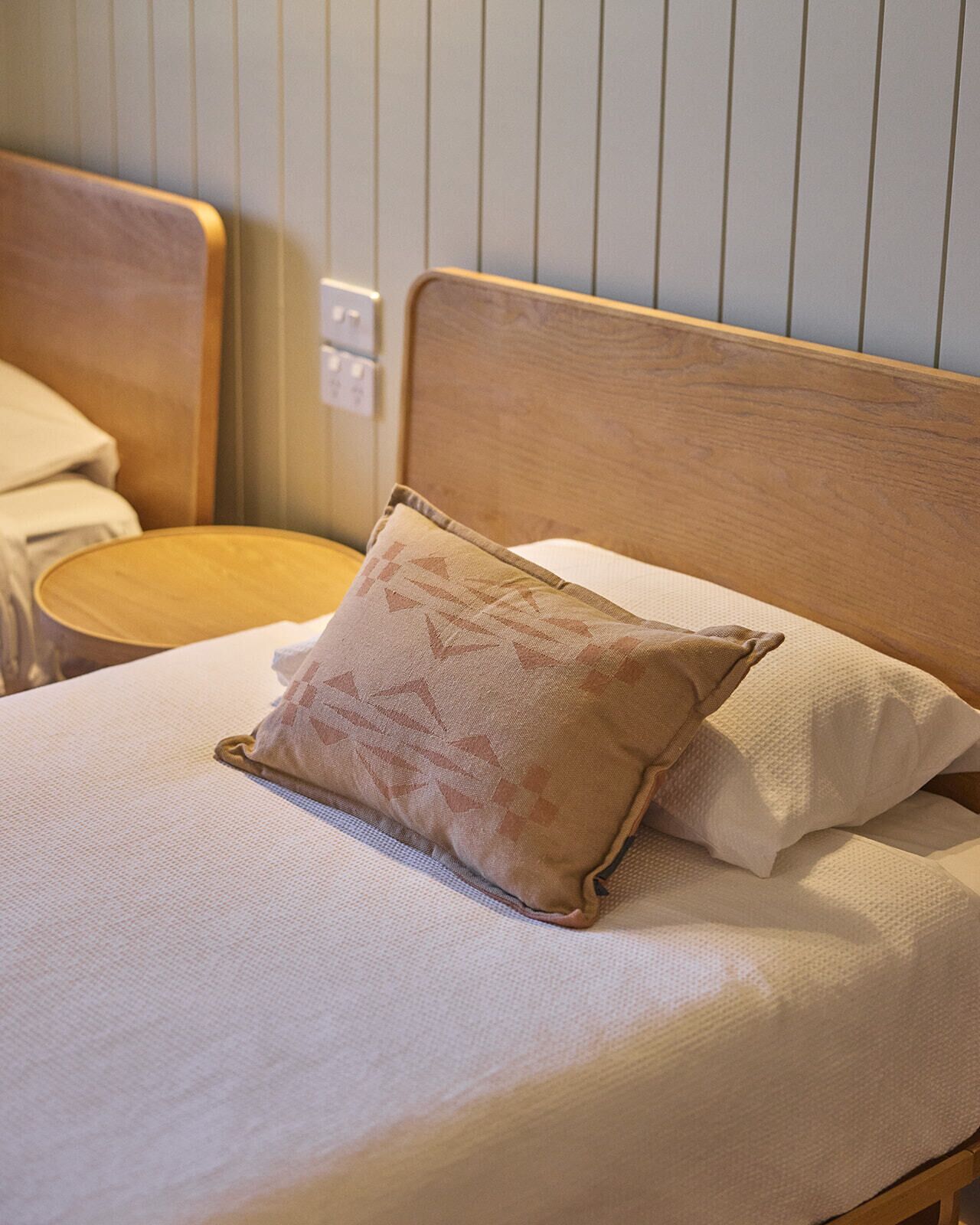 Ein ordentlich bezogenes Bett mit einem Holz-Kopfteil, einem beige gestreiften Kopfkissen und einem weißen Kopfkissen.