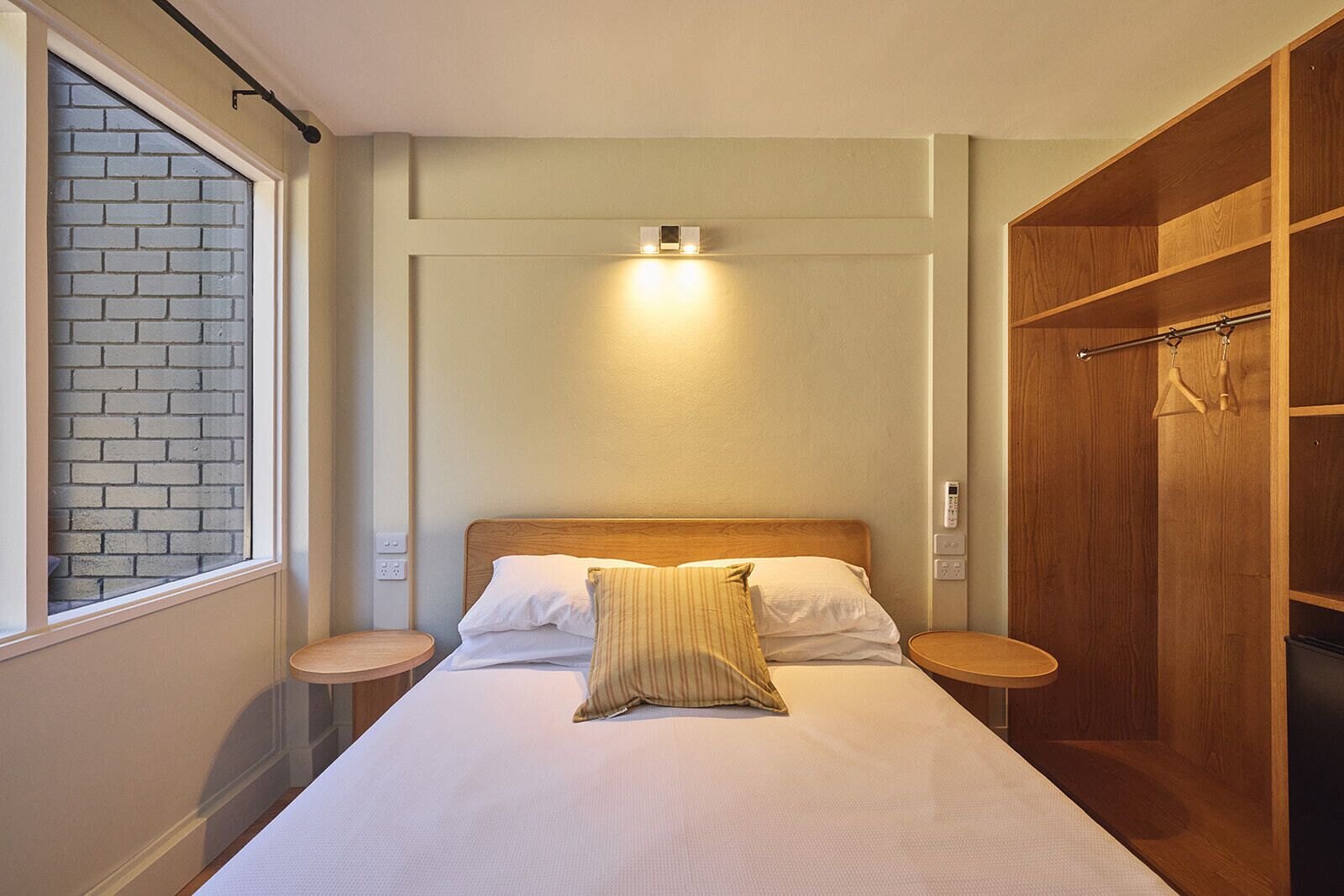 Une chambre à coucher comprenant un lit, des tables de chevet, une armoire en bois et une fenêtre donnant sur un mur de briques.