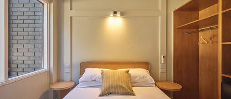 A bedroom with a bed, bedside tables, a wooden wardrobe, and a window with a brick wall view.